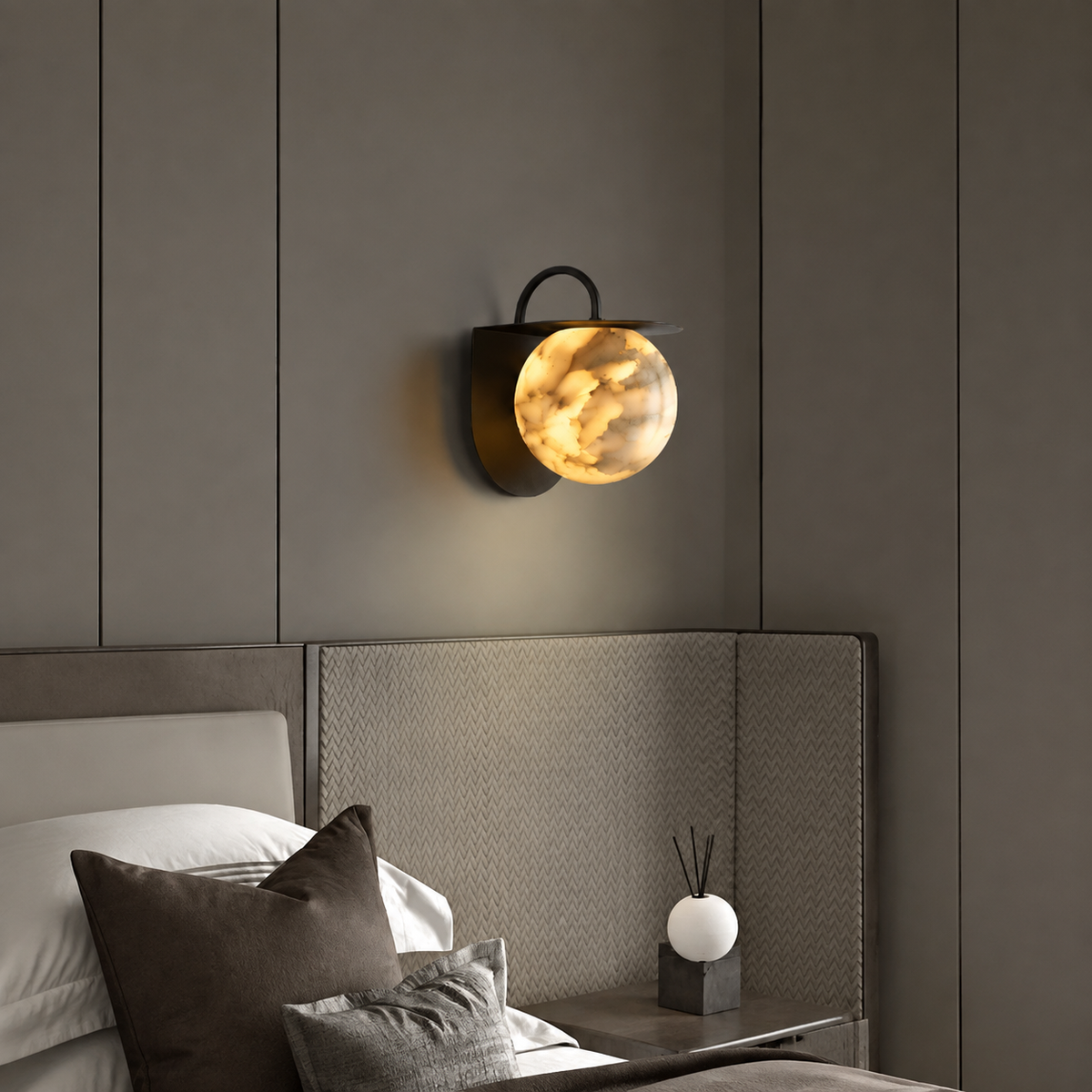 A modern wall-mounted sconce with a glowing amber glass globe and a dark metal frame, installed above a bed in a contemporary bedroom.