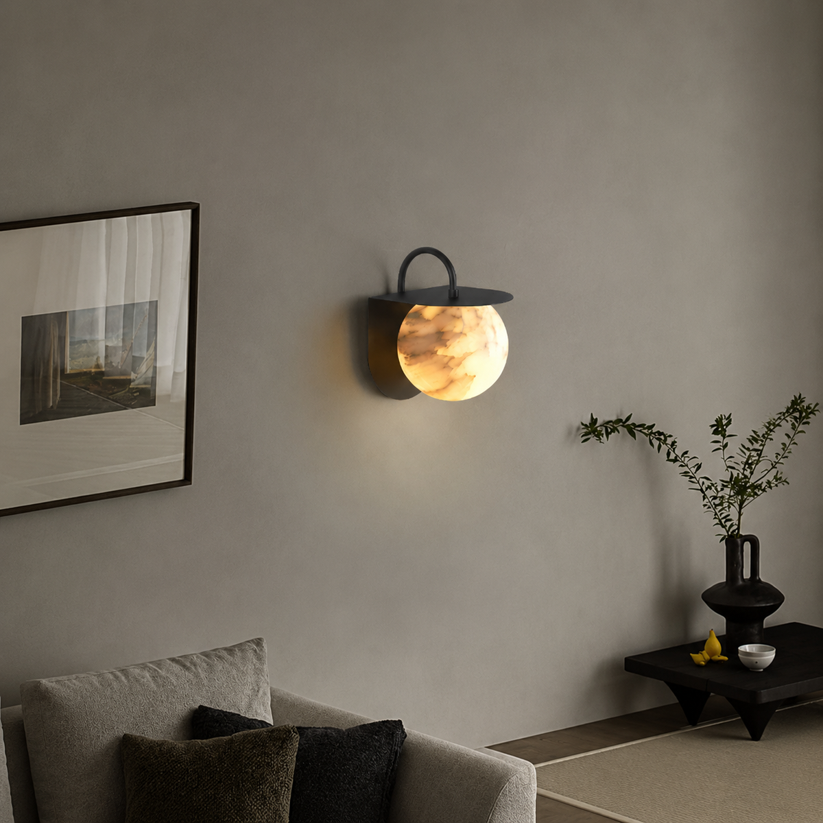 A modern wall-mounted sconce with a glowing oval glass shade and a dark metal top loop, installed on a gray wall in a styled interior with a sofa, framed art, and a small side table.