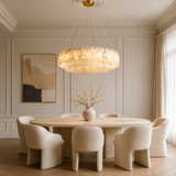 A modern dining room featuring a large circular pendant chandelier with a warm glowing cream marble-look shade, suspended over a round light wood dining table with six textured cream boucle chairs.