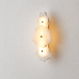 A modern wall sconce featuring three stacked, irregularly shaped white stone or resin discs that glow with warm yellow light from three small integrated bulbs, set against a neutral light gray background.