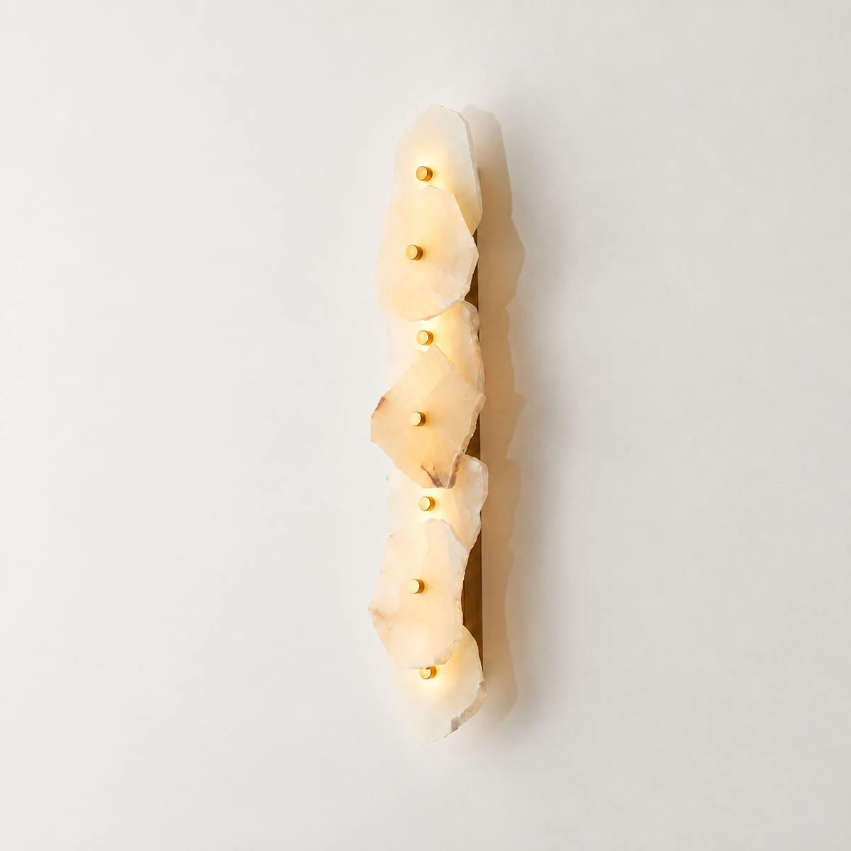 A vertical wall sconce featuring five stacked, irregular natural stone slices that glow warm white when lit, with small gold-toned bulbs set into the center of each stone. The fixture is shown against a clean, off-white background with a soft shadow.
