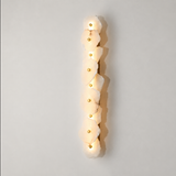 A modern vertical wall sconce featuring a column of irregular, organic-shaped frosted white stone or acrylic panels, each illuminated from within with a warm glow and accented by a small gold circular detail, set against a neutral light gray background.