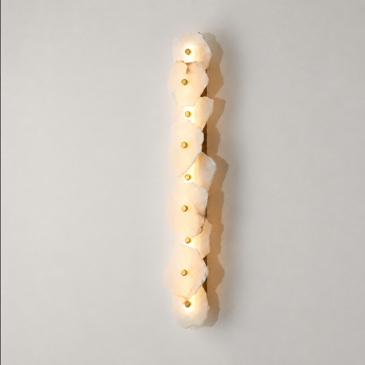 A modern vertical wall sconce featuring a column of irregular, organic-shaped frosted white stone or acrylic panels, each illuminated from within with a warm glow and accented by a small gold circular detail, set against a neutral light gray background.