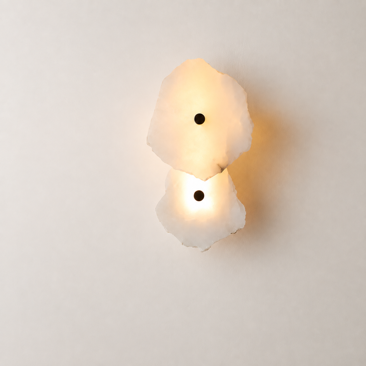 A modern two-light wall sconce featuring organic, irregularly shaped frosted white stone or glass shades that glow warm yellow when lit, with small black circular accents in the center of each light, mounted on a plain off-white wall.