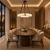A modern dining room featuring a large oval wooden dining table surrounded by eight upholstered chairs, under a large circular chandelier. The room has warm wood paneling, a textured beige wall, and large windows showing a sunset view.