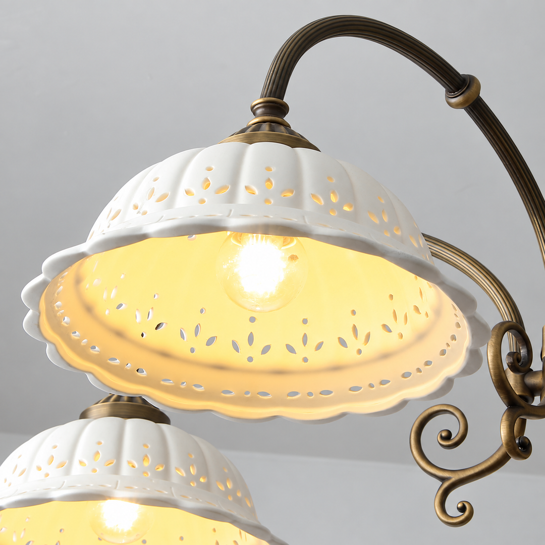 Chandelier close-up showing illuminated white fluted ceramic shades with decorative cutouts and curved aged brass arms against a gray background.