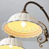 Chandelier close-up showing illuminated white fluted ceramic shades with decorative cutouts and curved aged brass arms against a gray background.