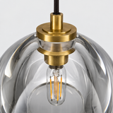 Close-up of a pendant light with a clear glass dome shade, brass metal fixture, and visible filament bulb against a light gray background.