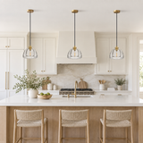 Three modern pendant lights with clear glass dome shades and gold accents hang over a white kitchen island in a bright, contemporary kitchen with white cabinets and wood accents.