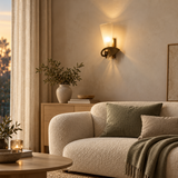 A warm brass wall sconce with a frosted glass shade mounted on a textured beige wall above a cream bouclé sofa in a cozy living room setting.