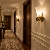 A warm, elegant hallway with dark wood doors, white wainscoting, a patterned runner rug, and two illuminated wall sconces mounted on the cream-colored walls.
