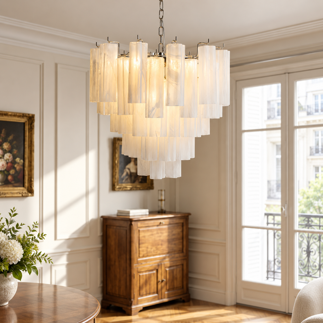 A modern multi-tier chandelier with vertical white fabric shades hangs in a bright, elegant room with white paneled walls, a wooden cabinet, and large French doors.