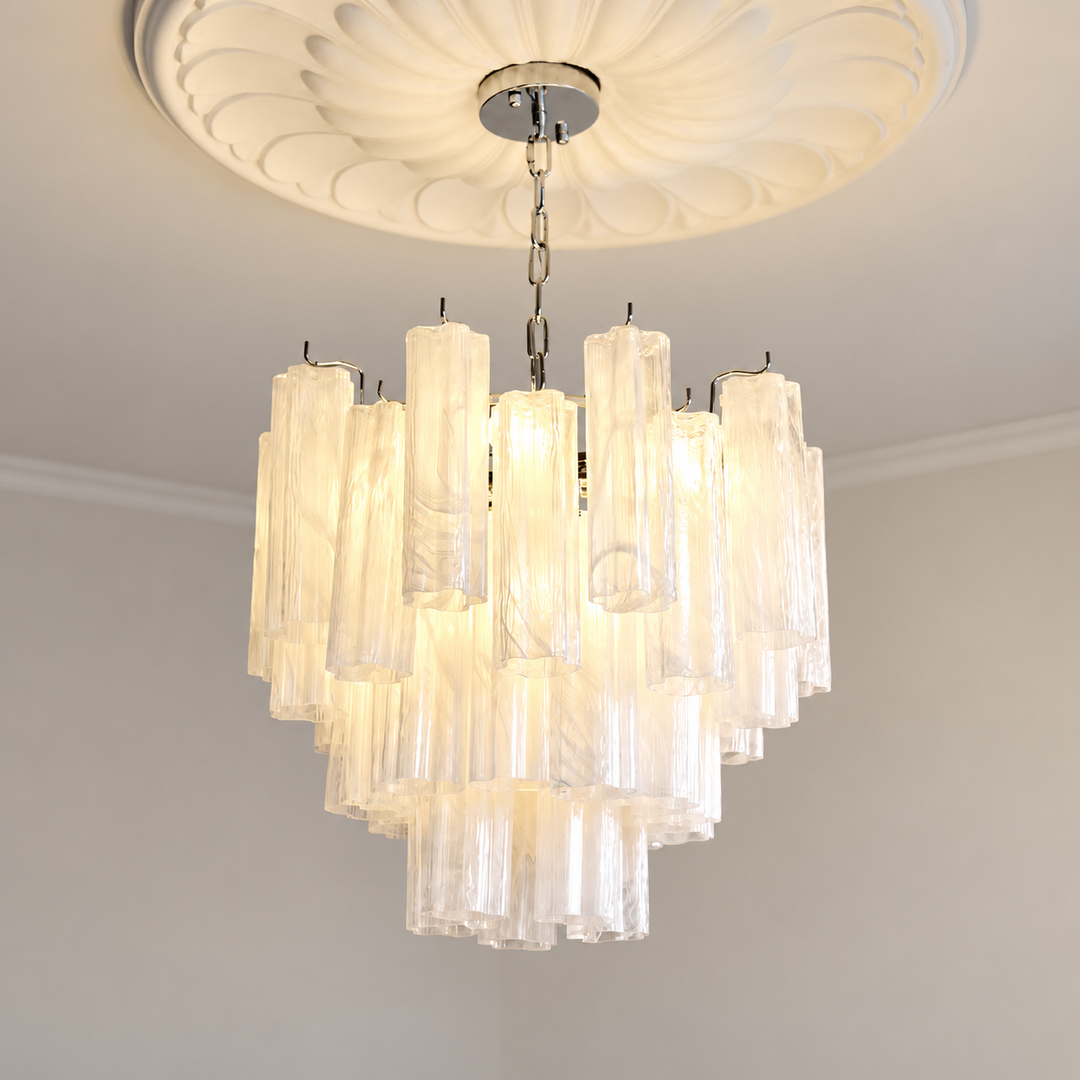 A modern multi-tier chandelier with clear textured glass cylindrical tubes, suspended from a silver ceiling canopy and ornate white ceiling medallion.