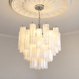 A modern multi-tier chandelier with clear textured glass cylindrical tubes, suspended from a silver ceiling canopy and ornate white ceiling medallion.