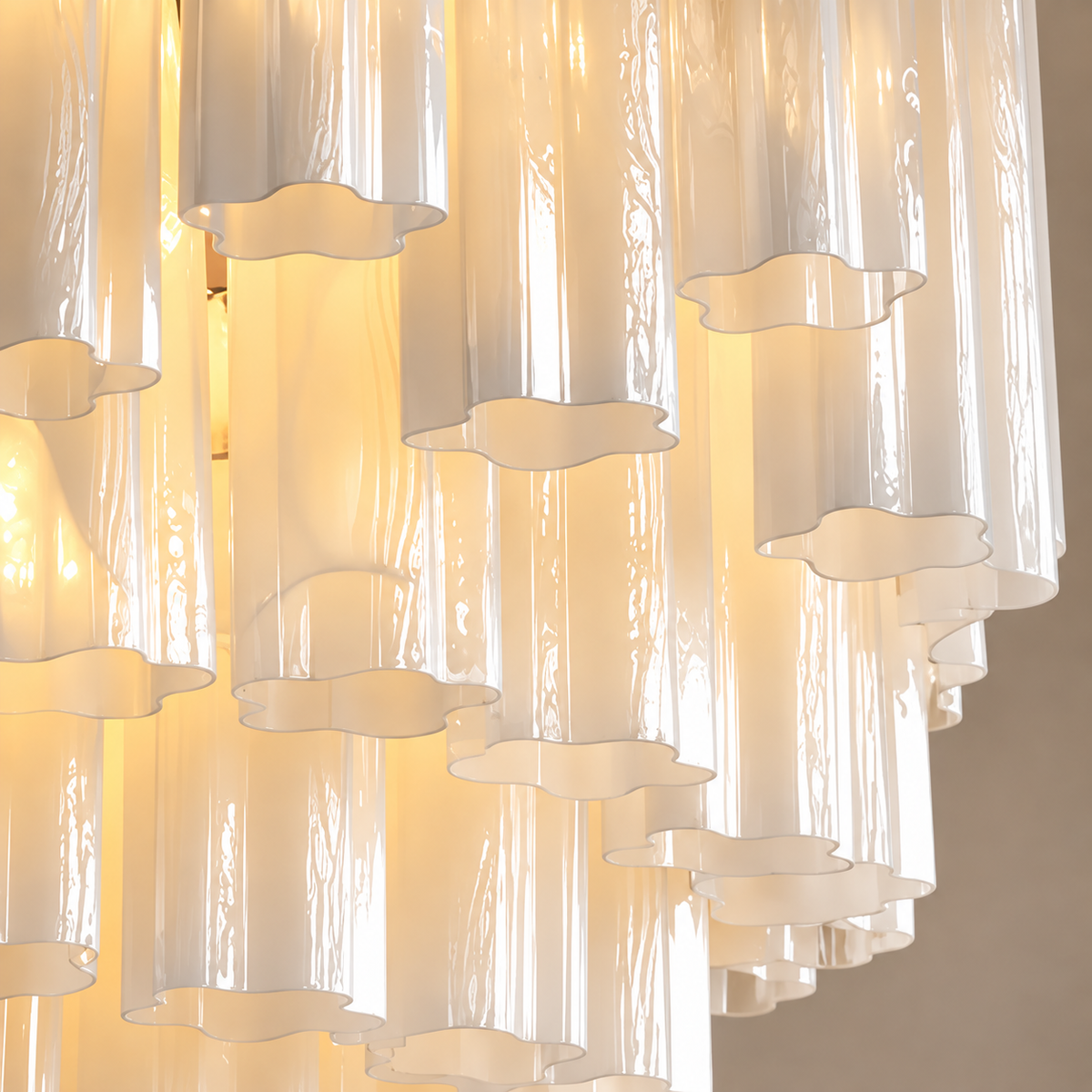 A close-up of a modern chandelier with multiple vertical frosted glass shades in varying sizes, layered with wavy, cloud-like cutouts, glowing with a warm yellow light against a neutral beige background.