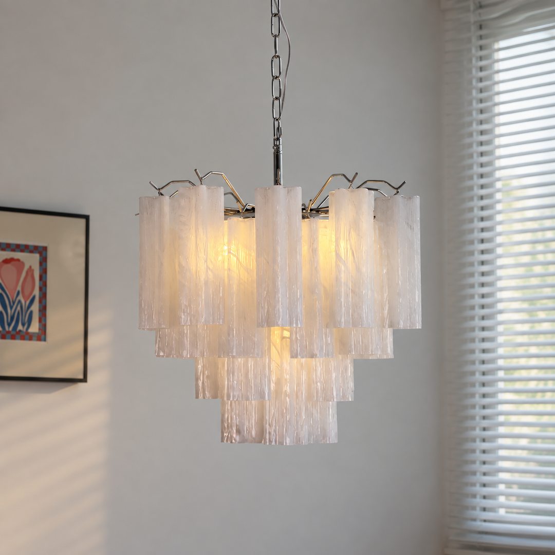 A modern multi-tier chandelier with textured, translucent white glass cylindrical shades and a silver metal frame, hanging in a room with a window and wall art visible in the background.