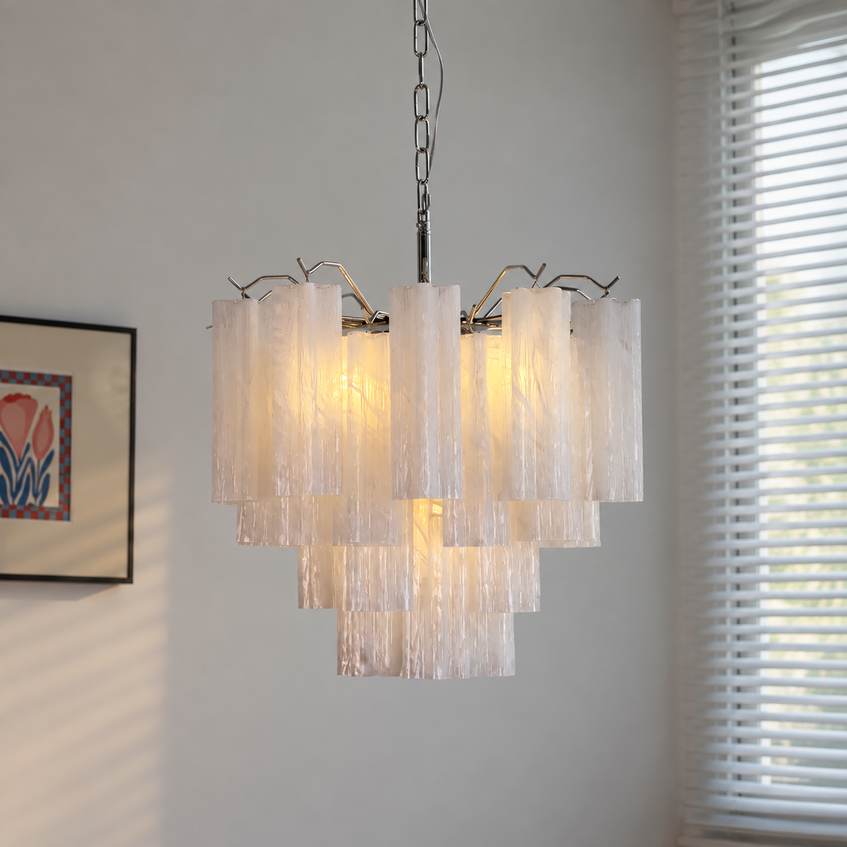 A modern multi-tier chandelier with textured, translucent white glass cylindrical shades and a silver metal frame, hanging in a room with a window and wall art visible in the background.