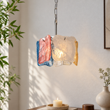 A modern pendant light with three textured, hand-blown glass panels in blue, pink, and clear/white, illuminated with a warm light. The light hangs from a silver chain in a styled room setting with a candle on a table below and a plant on the left.