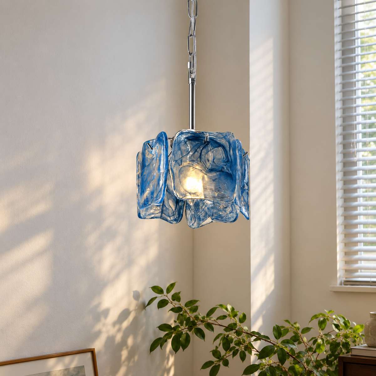 A blue textured glass pendant light hanging in a sunlit room corner, with a warm bulb glowing inside. The light fixture has a wavy, organic glass shade and a silver metal chain and rod.