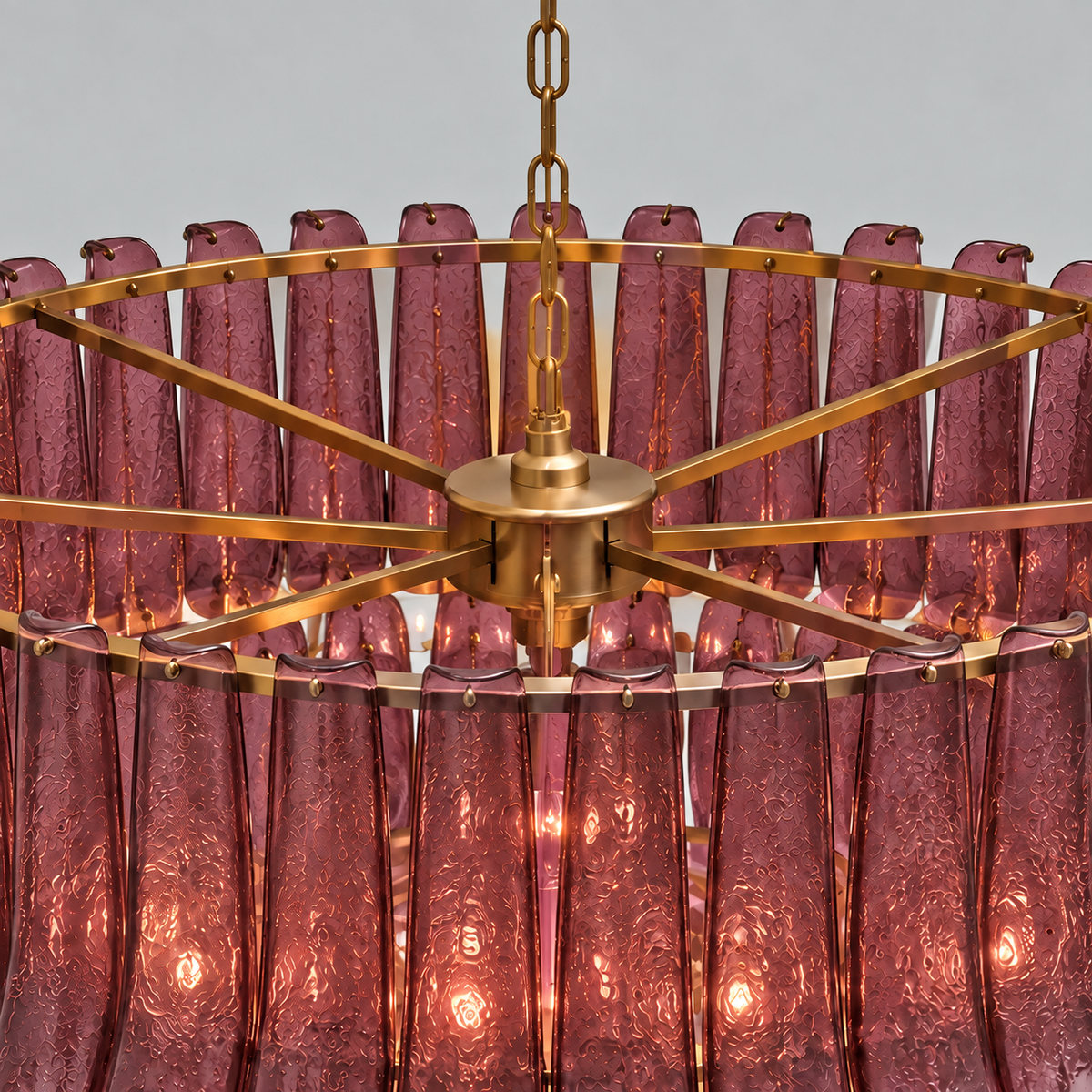 A close-up of a modern chandelier featuring a circular frame made of brushed brass/gold metal and numerous textured, translucent pink glass shades hanging from the center, illuminated with warm light.