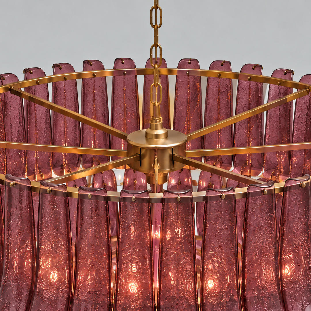A close-up of a modern chandelier featuring a circular frame made of brushed brass/gold metal and numerous textured, translucent pink glass shades hanging from the center, illuminated with warm light.