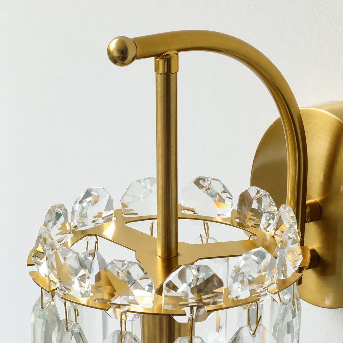 Close-up of a gold wall sconce with a curved arm, central post, and a ring of clear hanging crystal prisms against a plain light background.