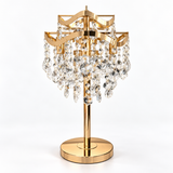 A gold-finished table lamp with a tiered crystal chandelier design, featuring clear faceted hanging prisms on a round base, photographed against a seamless white background.