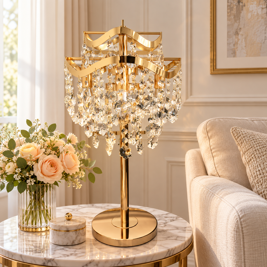 A luxury table lamp with a polished gold-finished frame and cascading clear crystal prisms, displayed on a marble side table in a beautifully decorated living room next to a sofa and a vase of roses.