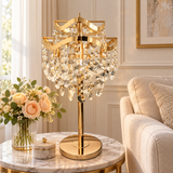 A luxury table lamp with a polished gold-finished frame and cascading clear crystal prisms, displayed on a marble side table in a beautifully decorated living room next to a sofa and a vase of roses.