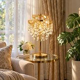 A luxurious gold table lamp with cascading crystal prisms stands on a round glass side table in a beautifully decorated living room with beige curtains, a sofa, and a potted monstera plant.