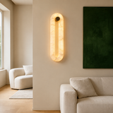 A modern wall-mounted sconce with a warm glowing oval frame made of light marble, installed on a beige wall in a minimalist living room setting.