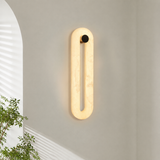 A modern vertical oval wall sconce with a warm glowing cream-colored marble-like frame and a small black circular accent at the top, mounted on a light gray wall in a styled interior setting with a plant and arched window visible.
