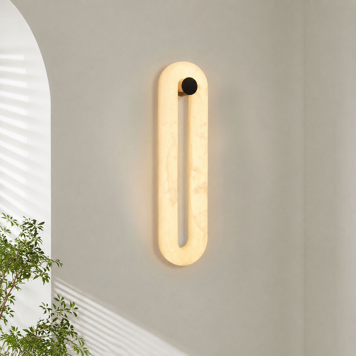 A modern vertical oval wall sconce with a warm glowing cream-colored marble-like frame and a small black circular accent at the top, mounted on a light gray wall in a styled interior setting with a plant and arched window visible.