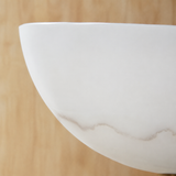 Close-up of a wall sconce's curved white Spanish alabaster shade with soft gray veining, set against a light wood background.