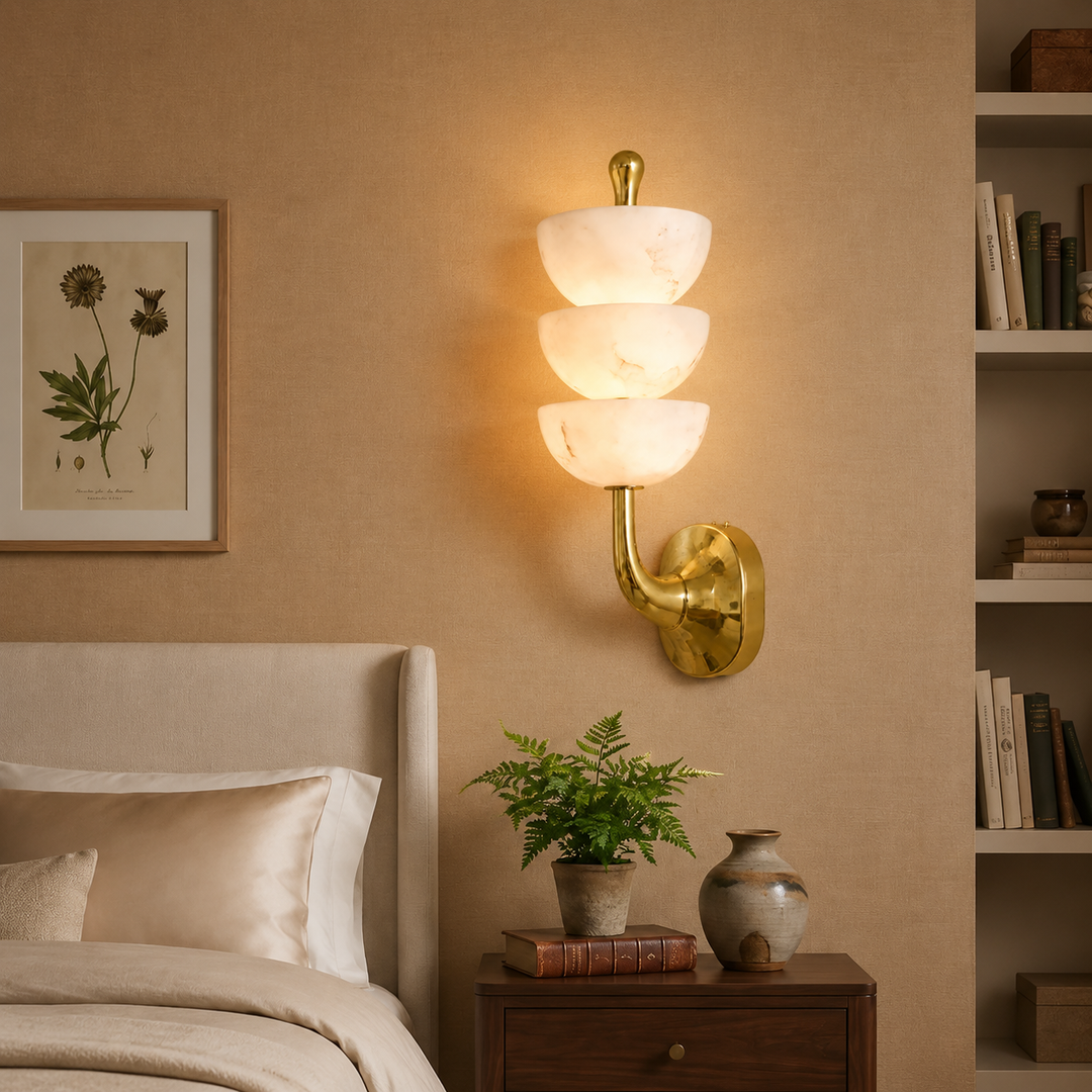 A wall-mounted three-tier sconce with white marble-look glass shades and a polished gold metal base, illuminated in a warm light on a beige bedroom wall above a wooden nightstand.