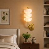 A wall-mounted three-tier sconce with white marble-look glass shades and a polished gold metal base, illuminated in a warm light on a beige bedroom wall above a wooden nightstand.