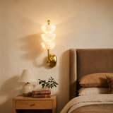 A modern wall-mounted sconce with three stacked frosted glass flower-like shades and a polished gold metal base, illuminated in a warm light on a beige bedroom wall above a wooden nightstand.