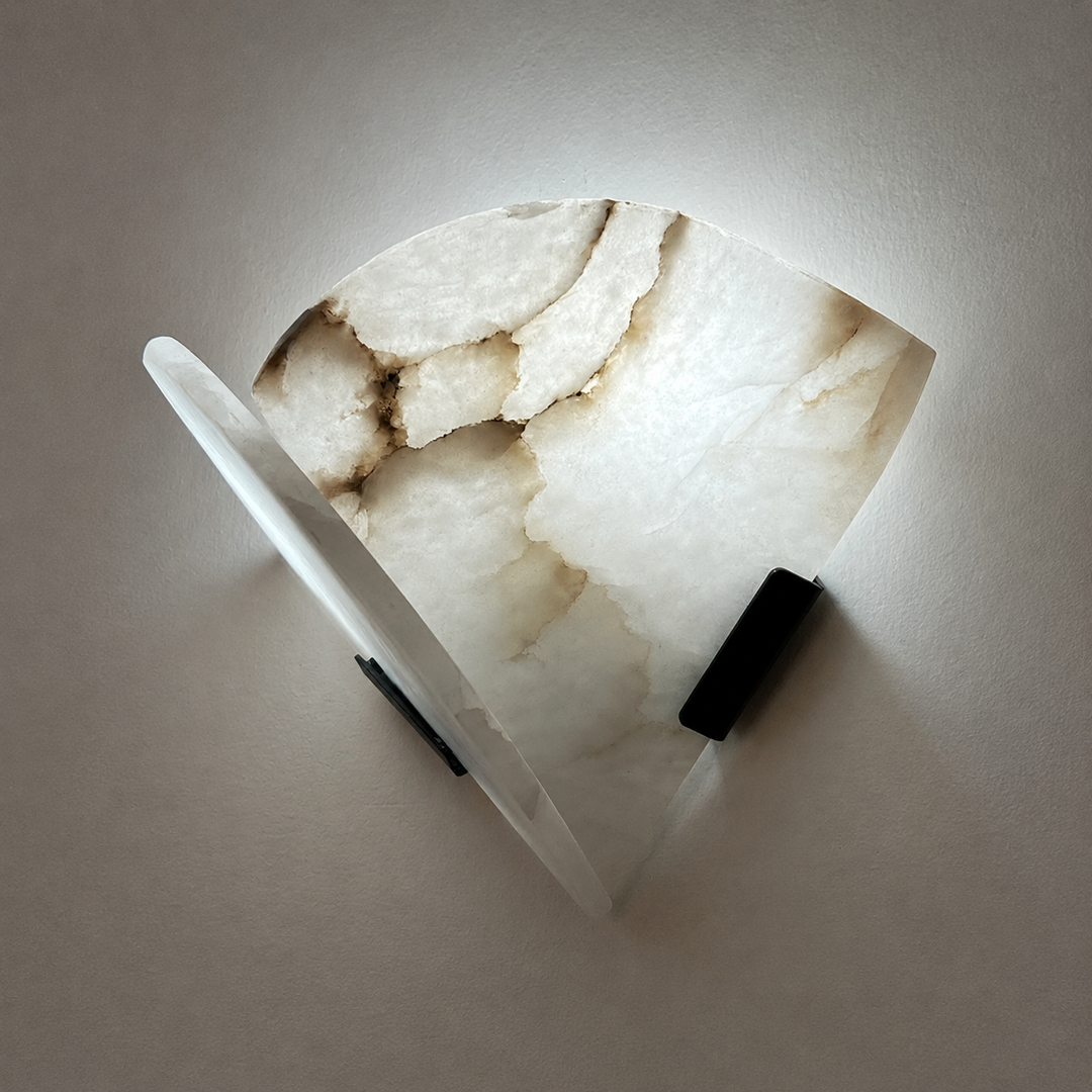 Angled view of a wall sconce shows layered white alabaster panels with beige veining, black supports and light against a plain wall.