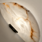 Close-up of a wall sconce shows an illuminated curved alabaster panel with amber-brown veining and a black iron support.