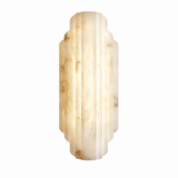 A modern wall sconce light fixture made of vertically fluted cream and beige onyx or marble, glowing with a warm light against a solid white background.