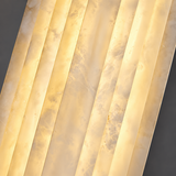 Close-up of a natural alabaster wall sconce with warm illumination revealing cream-colored veining and parallel ridges against a dark gray background.
