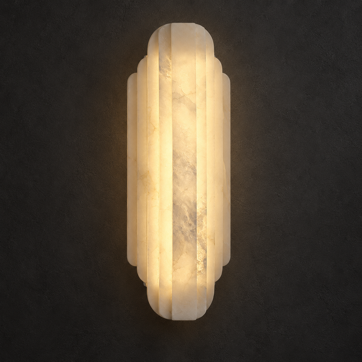 A vertical wall sconce made of warm white marble with a ribbed, tiered geometric design, glowing with a soft warm light against a dark textured wall.