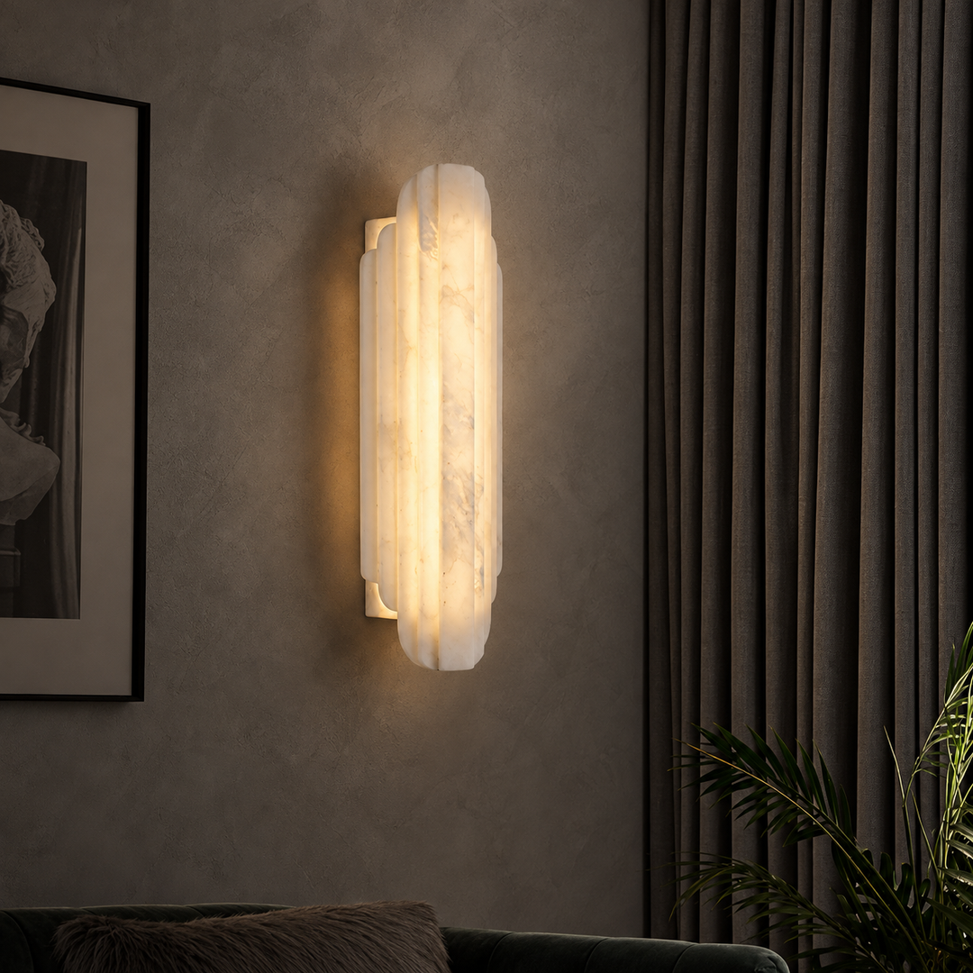 A vertical wall sconce with a warm white marble-look finish, mounted on a textured gray wall in a styled interior setting with curtains, a framed print, and a plant.