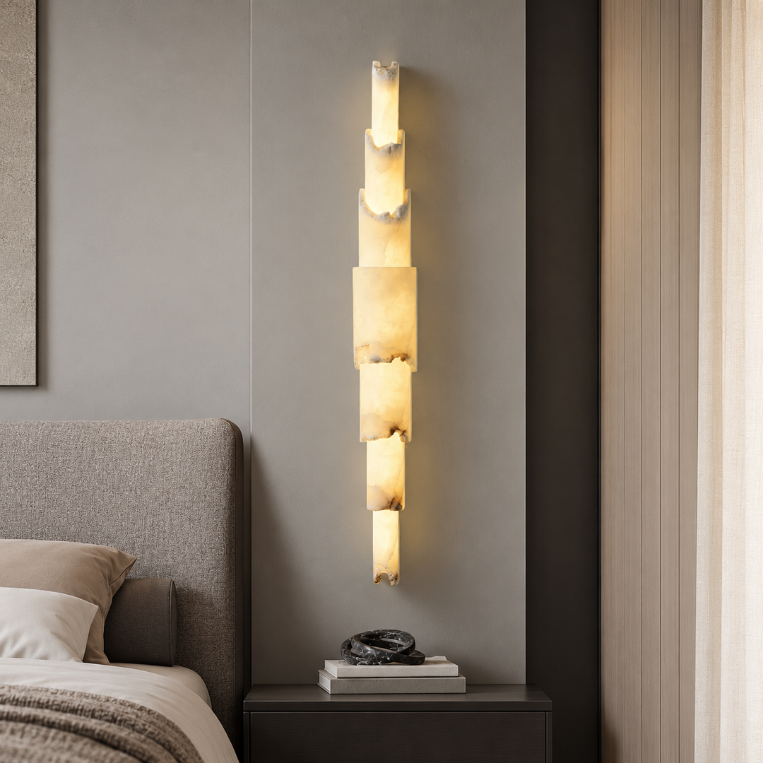 A modern vertical wall sconce with stacked rectangular illuminated stone or acrylic panels, mounted on a gray bedroom wall above a dark nightstand.