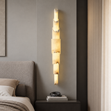 A modern vertical wall sconce with stacked rectangular illuminated stone or acrylic panels, mounted on a gray bedroom wall above a dark nightstand.