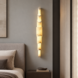 A modern vertical wall sconce with stacked rectangular stone-like panels glowing warm yellow light, mounted on a gray bedroom wall above a dark wood nightstand.