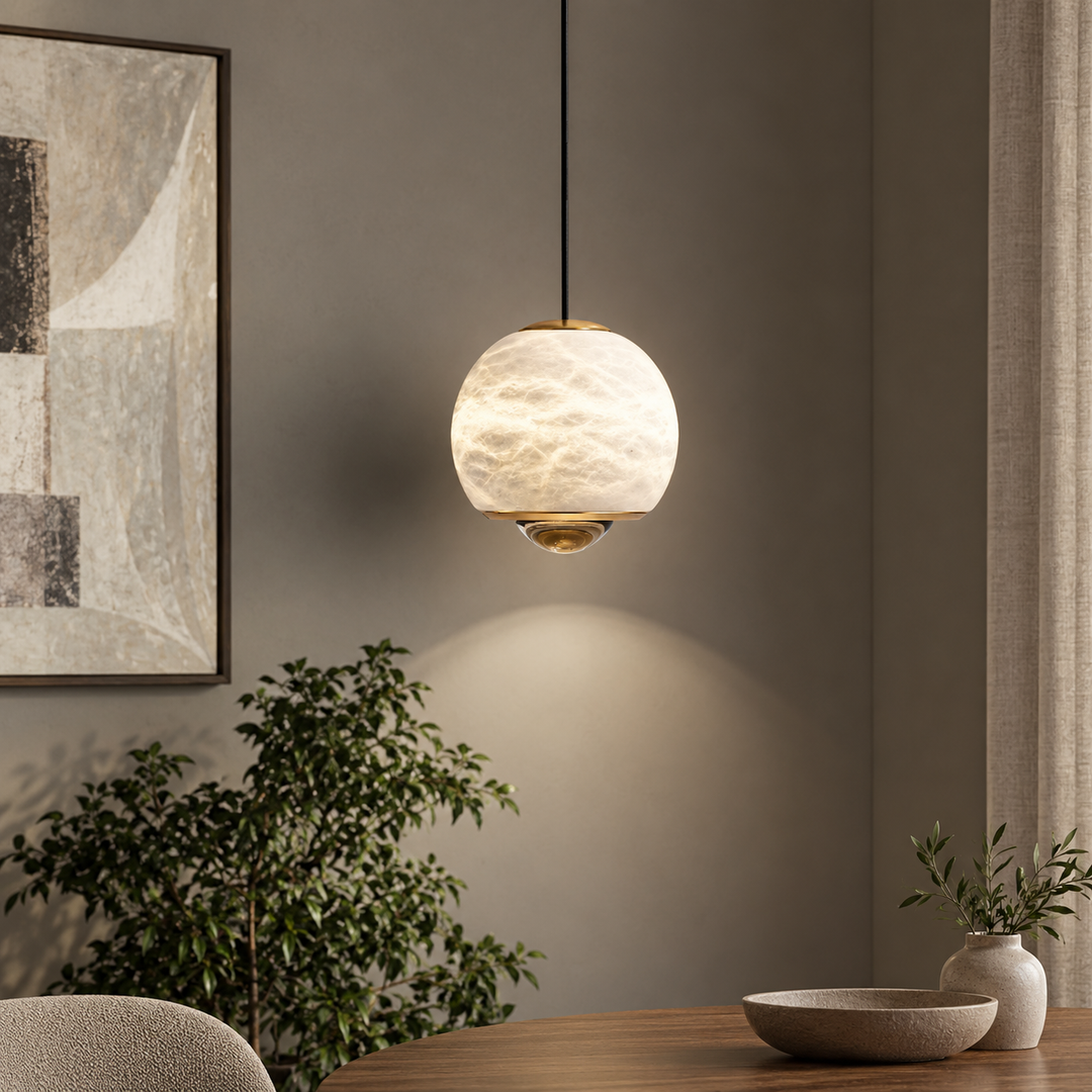A modern pendant light with a textured, frosted glass globe shade and brass hardware, hanging in a styled dining room setting with a wooden table, bowl, and potted plant.