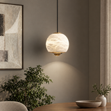A modern pendant light with a textured, frosted glass globe shade and brass hardware, hanging in a styled dining room setting with a wooden table, bowl, and potted plant.