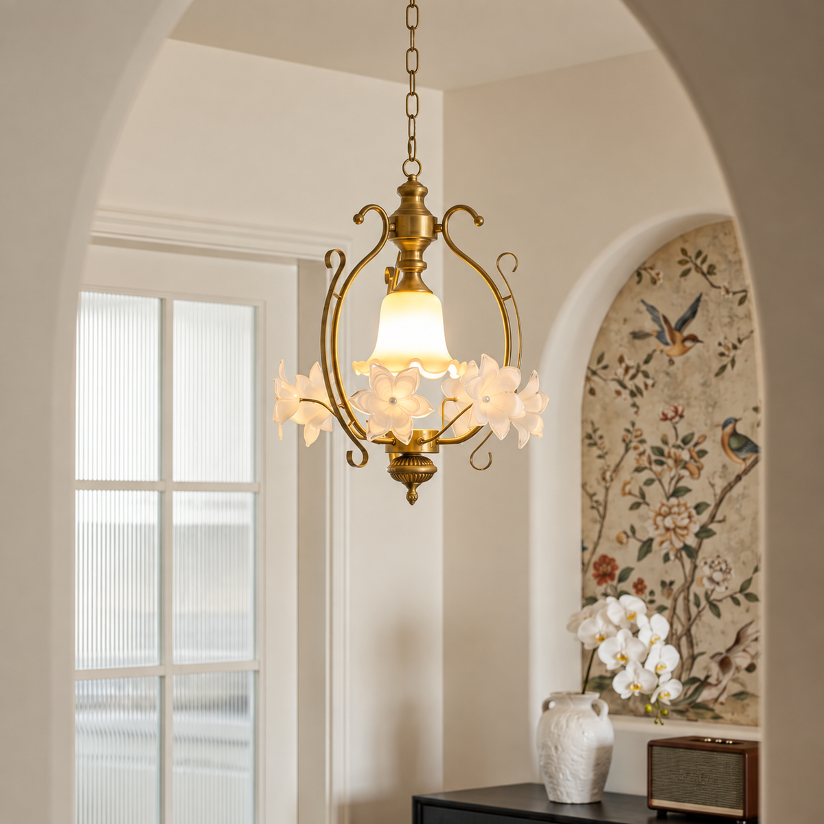 Gold-tone pendant light with cream glass flowers glowing in a bright room with an arched doorway and floral wallpaper.