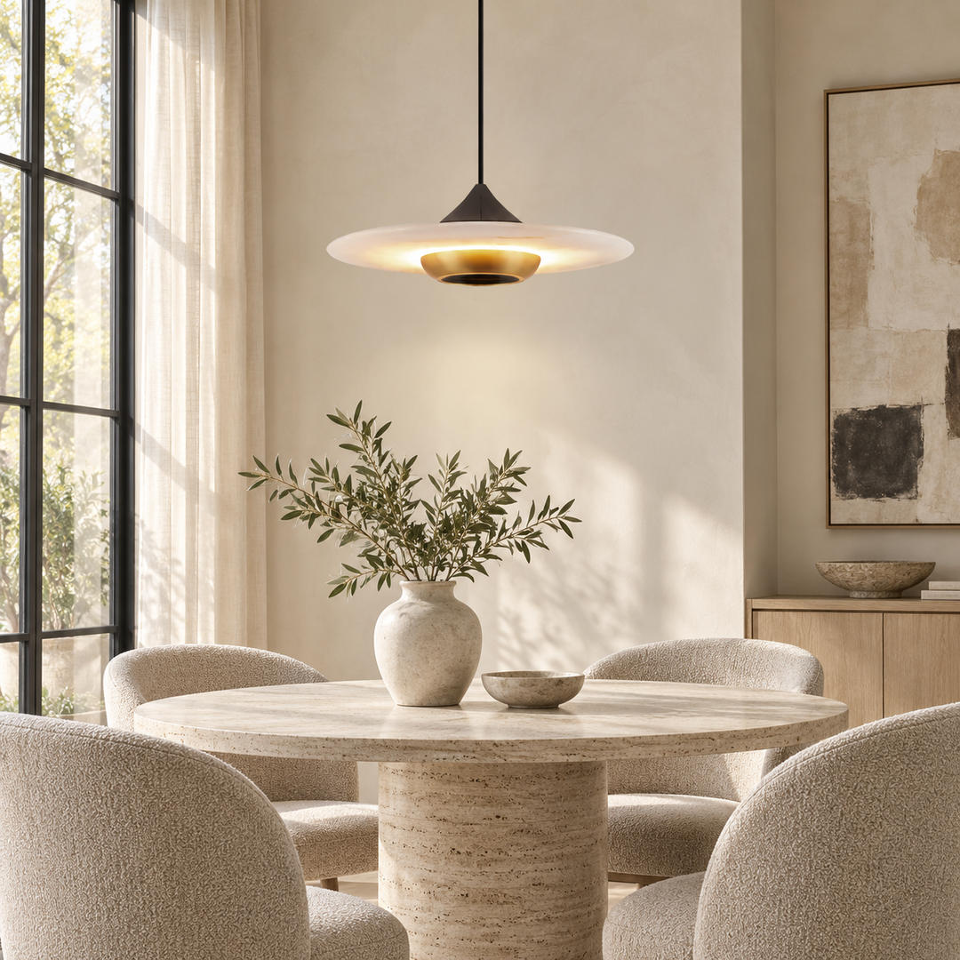 A modern pendant light with a wide white disc shade and a gold-toned glowing diffuser hangs over a round dining table in a softly lit neutral-toned dining room with upholstered chairs and a vase of greenery.